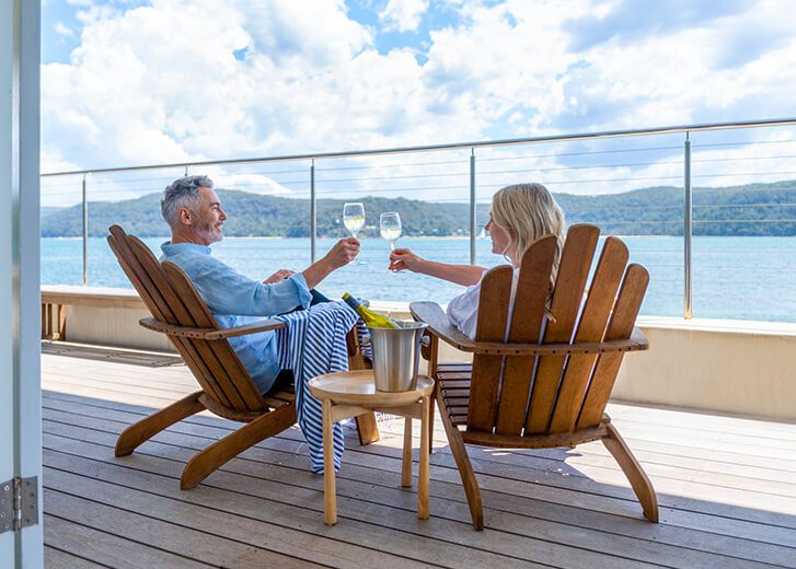 Mature couple drinking wine out on the deck. They are happy and smiling sitting in deck chairs. The sea is in the background